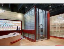 Moderne Apotheke mit geschwungenem Holz-Informationsschalter im Vordergrund, einer Wand mit Glasbausteinen auf der rechten Seite und einem automatisierten Medikamentenlager in einem verglasten Schrank. Im Hintergrund ist eine Wand mit Begrüßungen in verschiedenen Sprachen und digitalen Anzeigen für Medikamente zu sehen.