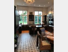 Eine geräumige, helle Küche mit dunklen Holzschränken, großen Fenstern mit Blick ins Grüne und einer zentralen Kücheninsel. Der Boden ist mit sechseckigen Fliesen ausgelegt, und ein Kronleuchter hängt von der Decke.