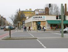 Straßenszene in einer urbanen Umgebung mit einer Kreuzung und einem Eckgebäude, das ein Second-Hand-Kaufhaus mit der Aufschrift „HUMANA“ zeigt. Mehrere Personen gehen über den Gehweg, während links Autos die Straße entlangfahren; im Hintergrund sind ältere Gebäude und Schilder von Geschäften sichtbar.