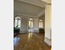 Innenraum eines historischen Wohnraums mit hohen, weiß gestrichenen Wänden und großen Rundbogenfenstern. Der Boden besteht aus glänzendem Parkett, und im Hintergrund führen Glastüren auf eine Terrasse mit Blick ins Grüne.