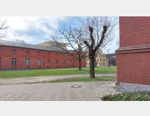 Backsteinbauten in einer parkähnlichen Umgebung mit gepflasterten Wegen, Rasenflächen und kahlen Bäumen. Im Hintergrund sind ein gelbes Gebäude und eine Glasfassade sichtbar.