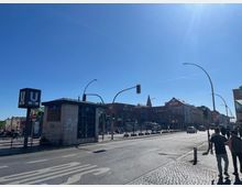 Straßenszene mit einer U-Bahn-Station der Linie U8 im Vordergrund, erkennbar durch das blaue "U"-Schild. Im Hintergrund sind mehrstöckige Gebäude, darunter ein Supermarkt mit einem roten "REWE"-Logo, sowie Bürgersteige, Verkehrsampeln und eine Hauptstraße bei sonnigem Wetter zu sehen.