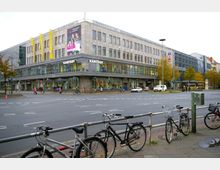 Mehrstöckiges Kaufhaus der Marke "Karstadt" mit großer Glasfassade im unteren Bereich und Steinstruktur im oberen Bereich, an einer belebten Straßenkreuzung. Im Vordergrund sind geparkte Fahrräder an einem Geländer zu sehen, während Fußgänger und Fahrzeuge die Straße nutzen.