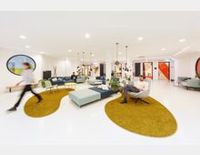 Heller, moderner Loungebereich mit weißen Wänden und hellen Bodenfliesen, ausgestaltet mit großen, organisch geformten Teppichen in Gelbgrün, blauen Sofas und grauen Sesseln. Mehrere Personen sitzen oder bewegen sich im Raum, Deckenlampen und grüne Pflanzen tragen zur Gestaltung bei.