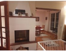 Wohnzimmer mit Kamin, Holzboden und angrenzendem Essbereich. Der Raum verfügt über helle Wände, große Fenster mit weißen Rahmen und eine Kinderschutzgitterumzäunung vor dem Kamin. Im Hintergrund steht ein Esstisch mit Stühlen.