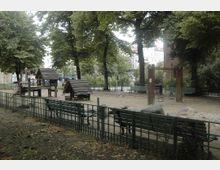 Städtischer Spielplatz mit kleinen Holzhäuschen, einer Schaukel und Sandfläche, umgeben von grünen Bänken, Bäumen und einem Metallzaun; im Hintergrund befinden sich Wohngebäude.