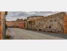 Gepflasterte Gasse, gesäumt von alten Stein- und Ziegelmauern, teilweise mit Efeu bewachsen. Im Hintergrund sind Dächer von Backsteinhäusern und ein wolkenbedeckter Himmel sichtbar.