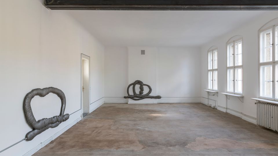 Ein leerer Raum mit weißen Wänden und mehreren hohen Fenstern auf der rechten Seite, die Tageslicht hereinlassen. Sichtbare Kunstinstallationen aus Metallrohren sind an den Wänden befestigt, der Boden ist roh und stellenweise fleckig.