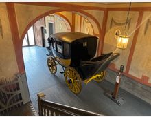 Eine historische Pferdekutsche mit schwarzen und gelben Elementen steht in einem Ausstellungsraum mit bemalten Wänden in klassischen Mustern. Der Raum hat Holzböden, eine hängende Laterne und einen offenen Eingang mit natürlichem Lichteinfall.