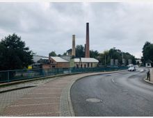 Straßenansicht mit einer gepflasterten Gehwegfläche und einer leicht geschwungenen Fahrbahn, die über eine Brücke mit grünen Metallgeländern führt. Im Hintergrund sind Industriebauten mit Ziegelfassaden und zwei hohen Schornsteinen zu sehen, umgeben von Bäumen unter bewölktem Himmel.