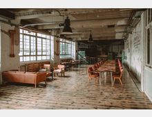 Industrieartig gestalteter Innenraum mit großen Fenstern, rohen Wänden und sichtbaren Leitungen an der Decke. Der Raum ist möbliert mit Ledersofas, Holzstühlen und einem langen Tisch mit roten Polsterstühlen auf einem Holzboden.
