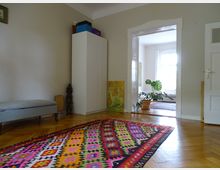 Ein helles Wohnzimmer mit Holzboden und einem bunten Teppich im Vordergrund. Im Hintergrund ist eine offene Tür zu einem angrenzenden Raum mit einem großen Fenster, einer Topfpflanze und einem weiteren Teppich zu sehen. Ein weißer Schrank, eine Bank und Dekorationsgegenstände befinden sich an den Wänden.