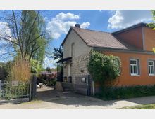 Ein Einfamilienhaus mit orangefarbener Fassade und teilweise sichtbarem Natursteinmauerwerk, umgeben von einem Garten mit Bäumen und Sträuchern. Im Vordergrund befinden sich ein metallisches Eingangstor und ein gepflasterter Weg.