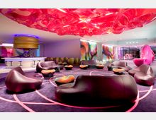 Eine moderne Lounge mit organisch geformten Ledersesseln und bunten Beistelltischen. Die Decke ist mit leuchtender, pink-transparenter Beleuchtung dekoriert, während abstrakte, farbige Wandmotive und eine helle Bar mit Tresenstühlen im Hintergrund zu sehen sind.