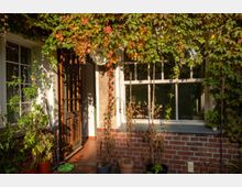 Eingangsbereich eines Hauses mit weißer Fassade und roten Ziegeln, großer Sprossenfensterfront und geöffneter Holztür. Rankpflanzen und Laub überwuchern die Veranda, davor stehen mehrere Topfpflanzen auf terrakottafarbenen Fliesen.