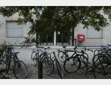 Ein moderner Gebäudeeingang mit Glas-Doppeltüren, flankiert von geschlossenen Fensterläden. Im Vordergrund stehen mehrere Fahrräder an einem Fahrradständer, teilweise umgeben von Bäumen.