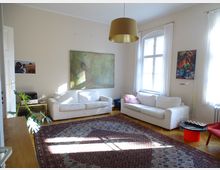 Helles Wohnzimmer mit zwei weißen Sofas, großen Fenstern mit weißen Vorhängen und einer goldenen Hängelampe. Ein großer Teppich bedeckt den Holzboden, an den Wänden hängen Kunstbilder, und links steht eine Zimmerpflanze neben einem kleinen Tisch mit einem Drucker.