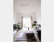 Helles Wohnzimmer mit modernen Möbeln, bestehend aus einem grauen Sofa, passendem Hocker und einem Fernseher auf einem niedrigen Regal. Der Raum hat helle Holzböden, große Fenster mit beigen Vorhängen, die auf einen Balkon führen, sowie Zimmerpflanzen als Dekoration.