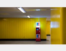 Geldautomat in einem Innenraum mit gelben Wandpaneelen und grauem Boden, beleuchtet von Deckenleuchten. Im Hintergrund befindet sich eine geschlossene Tür, die Teil der gelben Wand ist.