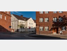 Straßenszene mit einer Kreuzung, umgeben von Gebäuden mit unterschiedlicher Architektur: links ein rotes Backsteingebäude, rechts ein modernes Backsteinhaus mit weißen Fenstern und orangefarbener Tür, sowie ein helles, klassisches Gebäude im Hintergrund. Die Straße ist gepflastert, mit einem Verkehrsschild und vereinzelten Bäumen am Straßenrand.