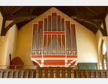 Innenraum einer Kirche mit einer großen, roten Orgel an der Wand, umgeben von glänzenden Pfeifen in verschiedenen Größen. Der Raum zeigt dunkles Holzgebälk an der Decke und rechts ein buntes Kirchenfenster.