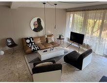 Wohnzimmer mit modernem Interieur, bestehend aus einem braunen Ledersofa, zwei schwarzen Sesselstühlen und einem weißen rechteckigen Couchtisch auf einem Teppich. Große Fenster mit transparenten Vorhängen bieten Blick auf einen Garten; an der Wand hängt ein rundes Kunstwerk.