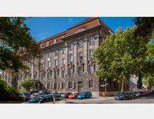 Ein historisches Gebäude mit massiver Steinfassade und großen rechteckigen Fenstern, gekrönt von einem roten Ziegeldach. Im Vordergrund stehen Autos am Straßenrand, und Bäume säumen die Straße unter einem klaren blauen Himmel.