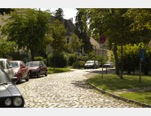 Kopfsteinpflasterstraße in einem Wohngebiet mit geparkten Autos entlang der Straße. Umgeben von Bäumen, Grünflächen und Wohnhäusern im Hintergrund.