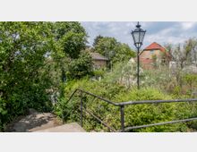 Treppenabgang mit Metallgeländer, umgeben von dichtem Grün und Bäumen, im Hintergrund Häuser mit roten Ziegeldächern und eine schwarze Straßenlaterne. Der Himmel ist teilweise bewölkt.