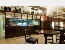 Innenraum eines Cafés oder einer Espresso-Bar mit dunklen Holzmöbeln, runden Tischen und Stühlen. Im Hintergrund befindet sich eine Theke mit einer offenen Küche, darüber Regale mit dekorativ aufgestellten Flaschen.