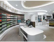 Innenansicht einer modernen Apotheke mit geschwungener Architektur. Die Wände sind mit Regalen voller Medikamente und Produkte bestückt, während im Vordergrund weiße Theken mit Kassensystemen und Kartenterminals sichtbar sind.