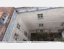 Blick in einen rechteckigen Innenhof umgeben von hellen Backsteinfassaden mit mehrfach unterteilten Fenstern. Im Hofbereich befinden sich ein verwitterter Boden mit Moosbewuchs und eine ungenutzte Metallkonstruktion.