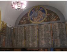 Innenraum einer Gedenkstätte mit einer gewölbten Decke. An der Wand befinden sich großflächige Tafeln mit langen Namenslisten, über denen ein Wandgemälde von Jesus mit ausgebreiteten Armen und floralen Ornamenten zu sehen ist. Ein Kronleuchter hängt von der Decke.