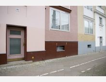 Fassade eines Wohngebäudes mit mehrfarbiger Putzstruktur in Rosa, Braun und Gelb. Sichtbar sind eine Holztür mit Glasfenster und goldfarbenem Griff, ein großes Fenster mit Vorhängen sowie ein kleines Kellerfenster. Der Gehweg besteht aus Kopfsteinpflaster und Betonplatten.