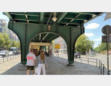 Ein grünes Stahlviadukt einer Hochbahn überspannt eine gepflasterte Straße mit Fußgängern. Im Hintergrund sind Bäume, Gebäude und Verkehrsschilder zu erkennen, die auf eine urbane Umgebung hinweisen.