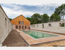 Ein Innenhof mit einem rechteckigen Swimmingpool, umgeben von einer Ziegelstein-Terrasse und einer weißen Steinmauer mit rechteckigen Öffnungen. Im Hintergrund steht ein Gebäude mit einer Fassade aus Holz und Ziegeln, dahinter befinden sich Bäume.
