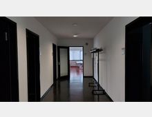 Ein schlicht gehaltener Flur mit weißen Wänden und dunkel glänzendem Boden. Rechts steht eine leere Garderobe, am Ende des Flurs führt eine geöffnete Glastür in einen Raum mit roten Bodenfliesen und Tischen an einem Fenster mit vertikalen Jalousien.