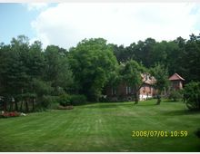 Großer Garten mit gepflegtem Rasen, umgeben von Bäumen und Blumenbeeten. Im Hintergrund steht ein Ziegelhaus mit roten Dachziegeln und Fenstern mit Blumenkästen, daneben ein kleineres turmähnliches Gebäude.