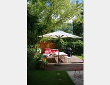 Ein gemütlicher Garten mit Holzterrasse, darauf eine Sitzgruppe mit weißen Möbeln und roten Kissen sowie ein weißer Sonnenschirm. Im Hintergrund befinden sich grüne Büsche und Bäume, darunter ein Holzzaun und ein Grill.