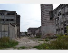 Ein verlassenes Industriegelände mit verfallenen Gebäuden und zerbrochenen Fenstern. Im Vordergrund sind überwuchertes Gras, eine zerstörte Betonfläche und ein großer Schornstein aus Backstein sichtbar.