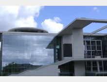 Modernes Gebäude mit großer Glasfassade und klaren, geometrischen Formen. Es verfügt über breite Treppen und ein flaches Dach, das teilweise über die Fassade hinaus ragt; der Himmel ist blau mit einigen Wolken.