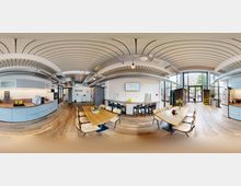 Großzügiger, moderner Gemeinschaftsraum mit Holzboden, einer offenen Küche auf der linken Seite, mehreren Holztischen mit Stühlen in der Mitte und großen Fensterfronten im Hintergrund, die viel Tageslicht hereinlassen. Sichtbare Deckenrohre und bogenförmige Deckenelemente betonen das industrielle Design.