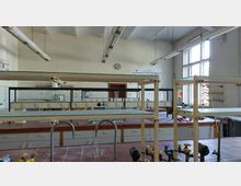 Ein chemisches Labor mit Arbeitsstationen aus roten Fliesen, mehreren Wasserhähnen und Regalen. Im Hintergrund befinden sich weiße Wandfliesen, ein Waschbecken, Poster und eine große Fensterfront mit einfallendem Tageslicht.