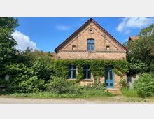 Backsteingebäude mit spitzem Giebeldach, blauen Fensterrahmen und einer blauen Tür, teilweise von grüner Kletterpflanze bewachsen. Es ist umgeben von Bäumen und Sträuchern unter einem blauen Himmel.