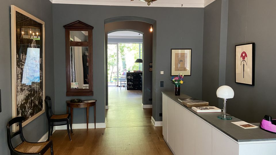 Innenansicht eines Flurs oder Empfangsbereichs mit grauen Wänden, Holzfußboden und Stuckdecke mit Kronleuchter. Rechts steht ein langer weißer Tresen mit Tischlampe und Bildern an der Wand, links hängen Kunstwerke und ein Spiegel; durch einen Rundbogen öffnet sich der Blick in einen hellen Raum mit Fensterfront zum Garten.