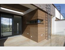 Moderne Dachterrasse mit Holzverkleidungen an den Wänden und einem gläsernen Geländer. Eine große Schiebetür führt von der Terrasse in den Innenraum, während eine Metallleiter an der Außenwand angebracht ist.