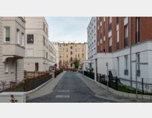 Schmale Wohnstraße mit modernen Mehrfamilienhäusern links und rechts, die weiß verputzt oder mit rotem Klinker verkleidet sind, im Hintergrund ein älteres sandfarbenes Gebäude. Die Straße ist asphaltiert und von niedrigen Zäunen sowie begrünten Flächen gesäumt.