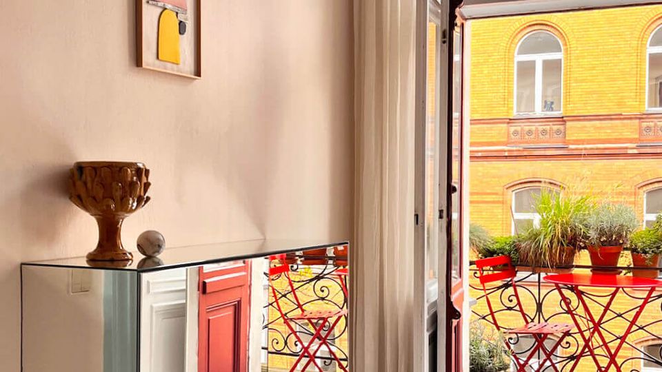 Bright apartment corner with white plank floors and ornate plaster ceiling molding, featuring a wall-mounted artwork and a mirrored console table. An open glass door leads to a small balcony with red bistro chairs and potted plants, overlooking a yellow brick building.