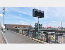 Eine Fußgängerbrücke mit Geländern führt zu einem Glasaufzug, der auf mehreren Ebenen zugänglich ist. Im Hintergrund sind die Mercedes-Benz Arena, ein rotes "Berlin"-Schild und Gebäude sichtbar; der Himmel ist leicht bewölkt.