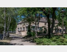 Einstöckiges Gebäude mit beigem Putz und Satteldach, umgeben von Bäumen und Gehölzen in einer parkähnlichen Umgebung. Ein gepflasterter Weg führt vorbei, und ein alter Straßenlaternenpfahl steht am rechten Bildrand.